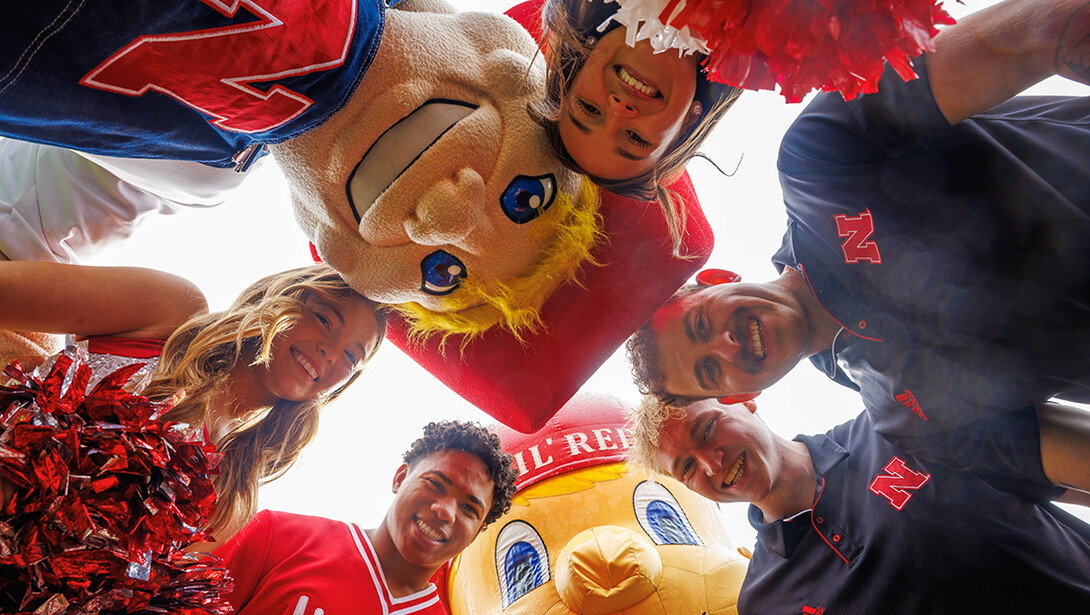 Aidan Kauth Fisher, a CEHS major who worked as Lil Red, Connor Hadaway, a Child Youth and Family Studies major who worked as Herbie in his undergrad, Amani Mfinanga, a senior Management major and Nebraska Spirit Squad member, Kylie Jones, a senior Marketing major and member of the Husker Scarlets and Grace Nab, a senior Nutritional Science and Dietetics major and member of the Nebraska Spirit Squad pose for a photo with Lil Red and Herbie on the turf in Memorial Stadium for their graduation.