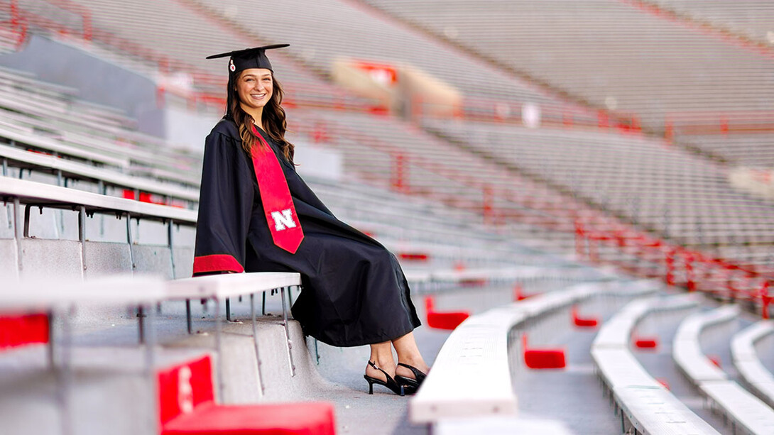 A girl wearing a graduation cap and gown sits on bleachers.