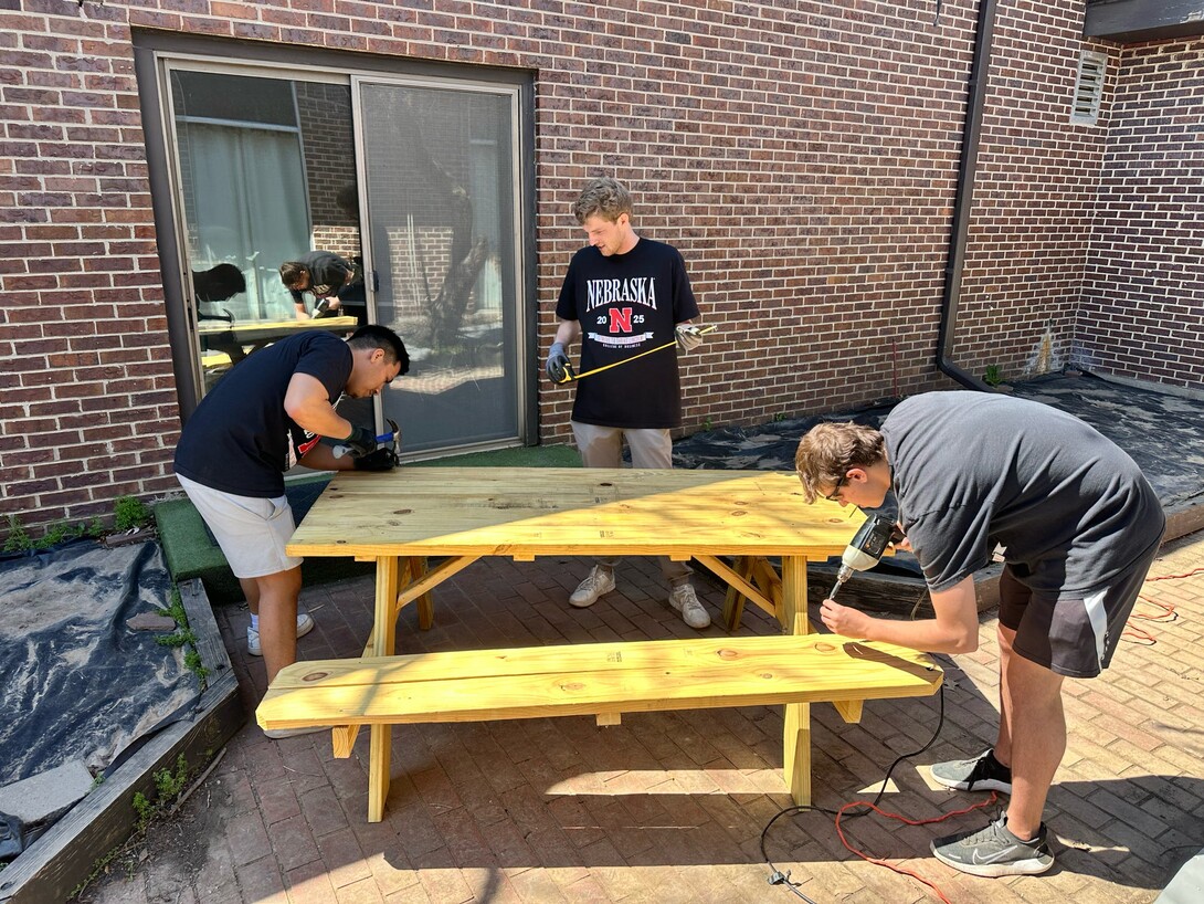 Three students in black Nebraska t-shirts constructing a yellow picnic table outside.