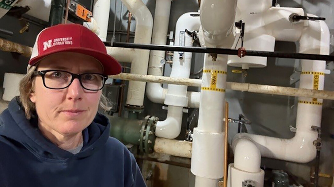 Beth Barry stands in a dark blue sweatshirt and a red Nebraska hat with rectangular glasses and short brown hair next to a system of white PVC pipes.