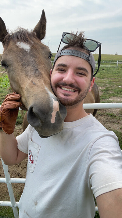 Baden Brumbaugh poses with a brown horse in a white t-shirt, backwards cap and sunglasses.