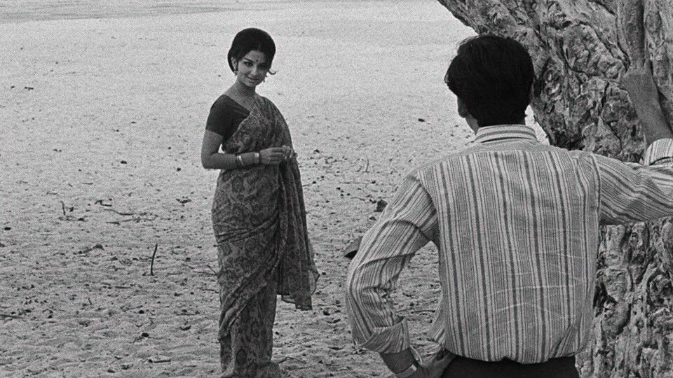 A black-and-white still of a brown woman in a saree stands looking back toward a man in a button-down shirt, who is leaning against a tree.