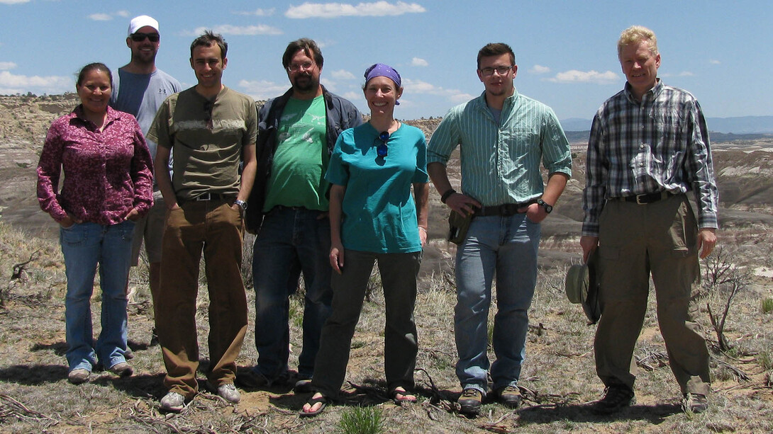 A group of seven people stand together for a group photo on dry land, a bright blue sky in the background.