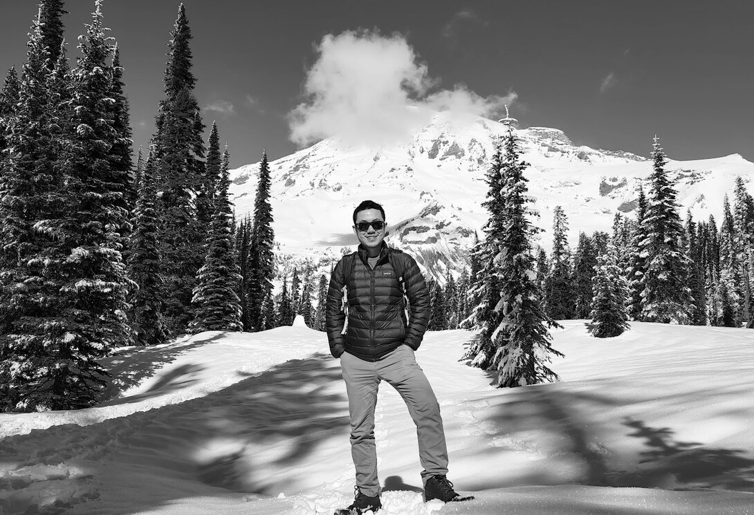 Liang Chen standing in snow with coniferous trees and a mountain peak in the background