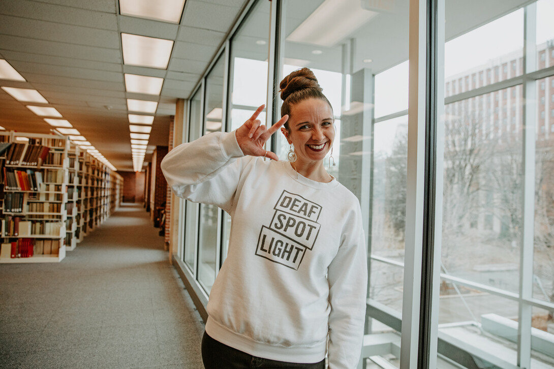 3rd-year PhD candidate Renca Dunn poses in Love Library, wearing a crew neck hoodie that reads "Deaf Spotlight" and signing "I love you."