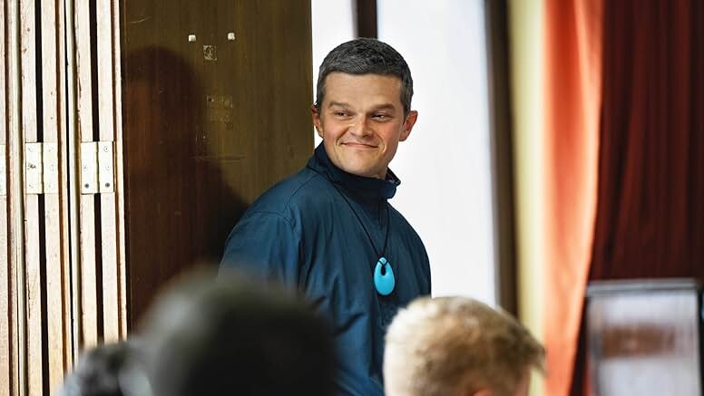 A white man with short black hair stands smiling in a blue tracksuit jacket.