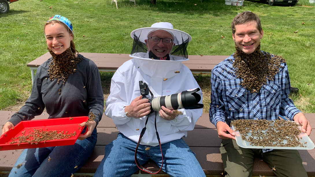 Photographer Craig Chandler in protective gear sitting on a bench between two students wearing beards made from live bees.