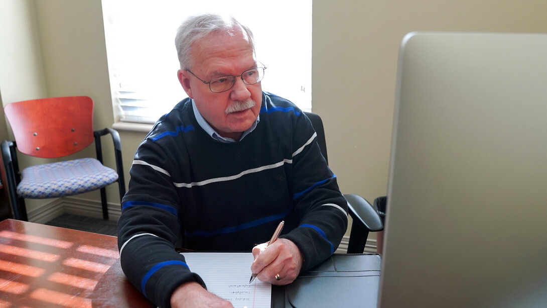 Michael Rubach, with grey hair and a mustache, sits in a blue sweater at his desk.