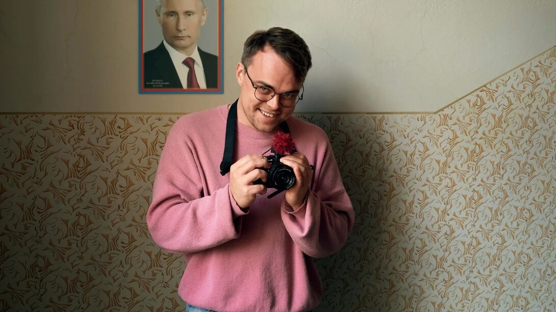 A teacher smiles as he holds a video camera with a portrait of Putin behind him.