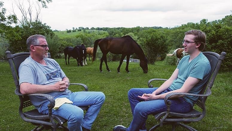 An older white man and younger white man, both in a t-shirt, jeans and glasses, sit across from each other in a green field outside, two horses grazing behind them.