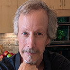 A white man with a long, thin face, grey hair and a grey goatie.
