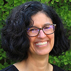 A woman with a dark, curly bob and black glasses smiles in front of a backdrop of green leaves.