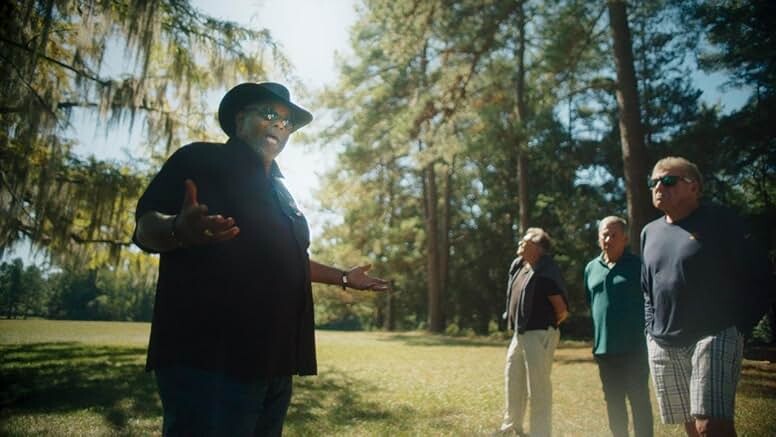 A tour guide, a Black man in a black shirt and black cowboy hat, stands in front of a small crowd outside surrounded by tall trees.