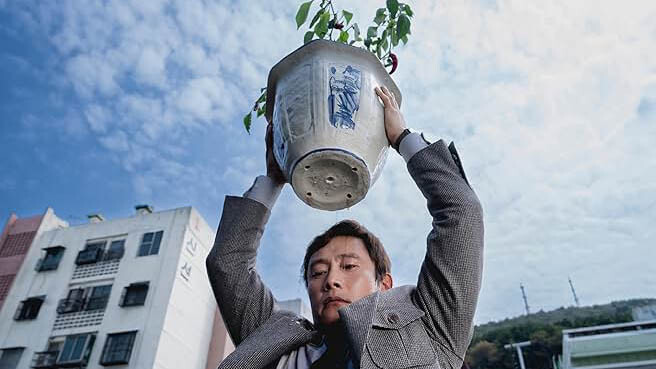 A man in a grey suit holds a potted chili plant above his head, getting ready to throw it to the ground.