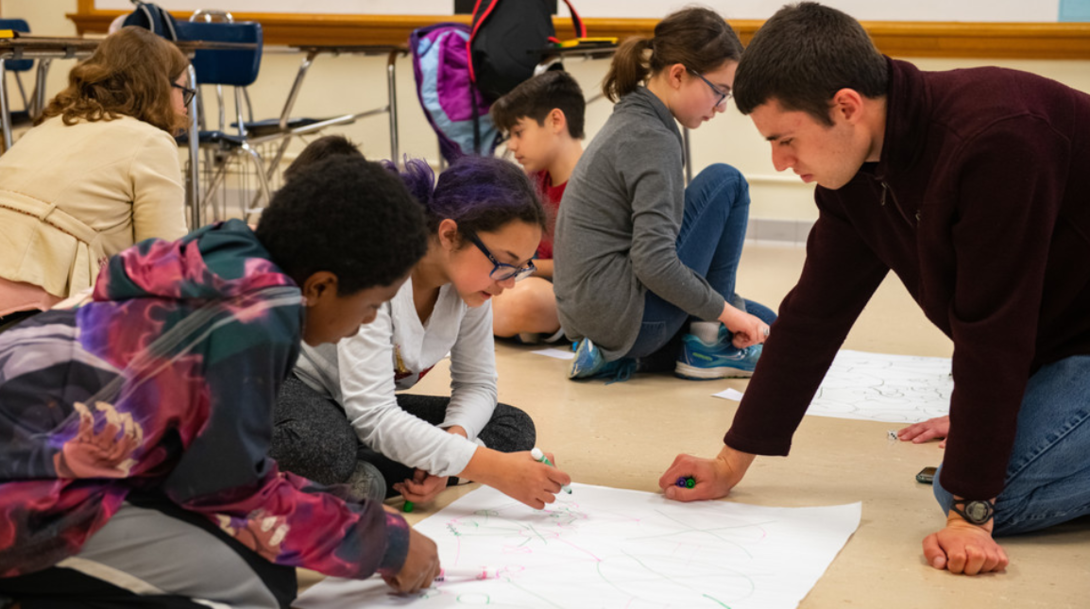 Worlds of Connections undergraduate mentor Luke Novak (right) works with a group of middle-school students to play a game demonstrating network science and the efficacy of vaccinations among community members.