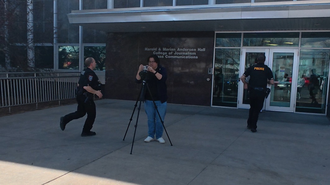Two people run into a building, passing a person standing behind a video camera.