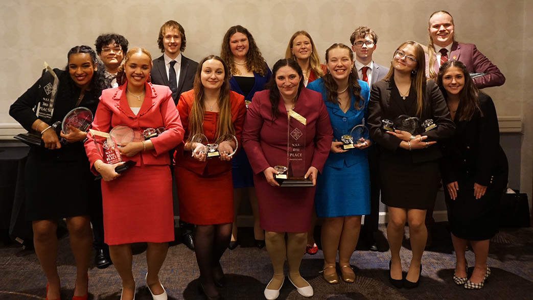 Thirteen students in formal clothing smile for a group photo with their awards.