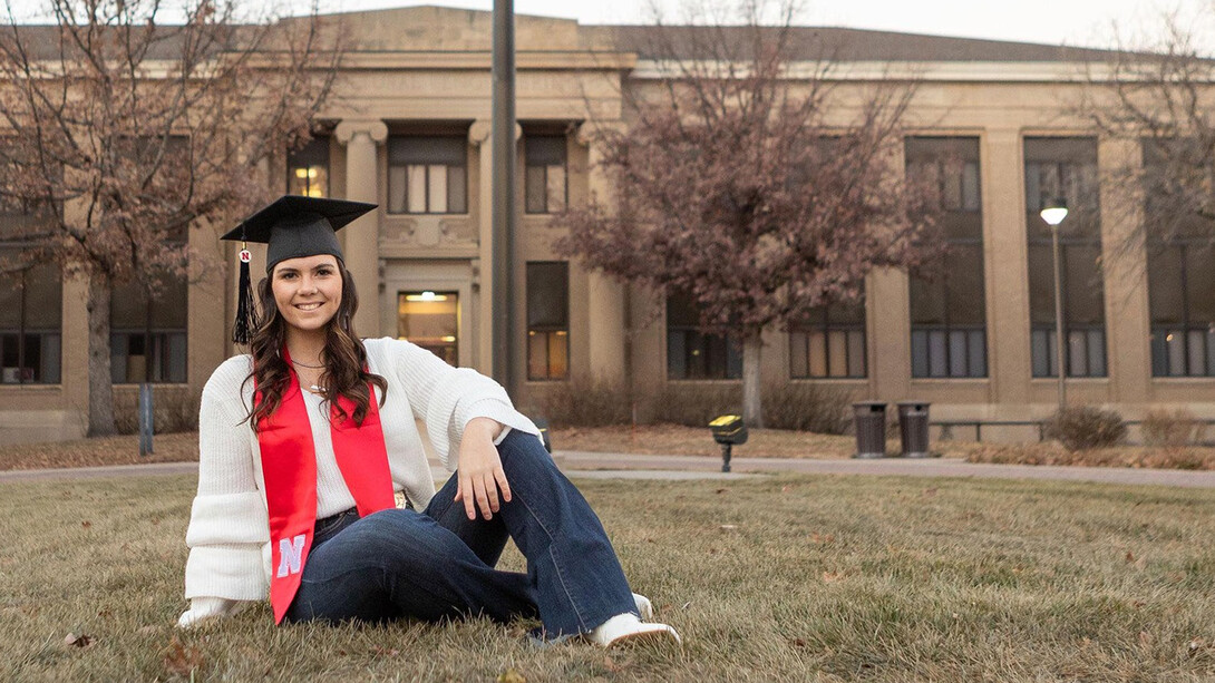Nebraska graduate Gracie Stout, wearing a mortar board and graduate stole, sits in the grass on the East Campus mall.
