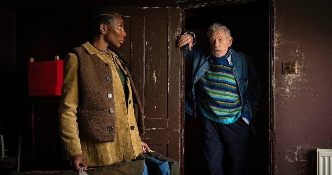 Michaela Cole, a dark-skinned Black woman, stands on the left in a tan and brown tunic, next to a doorway where Ian McKellan, an older white man, stands in a green and blue striped sweater.