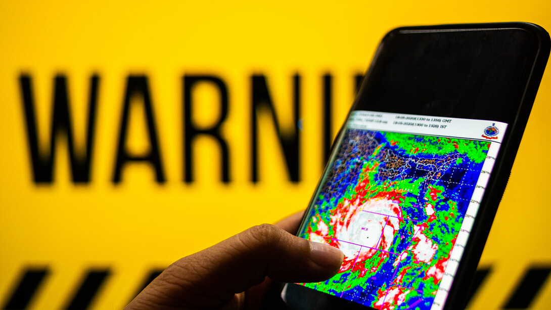 Photograph of a mobile phone with cyclone moving towards the coast shown. The background is yellow with warning sign.