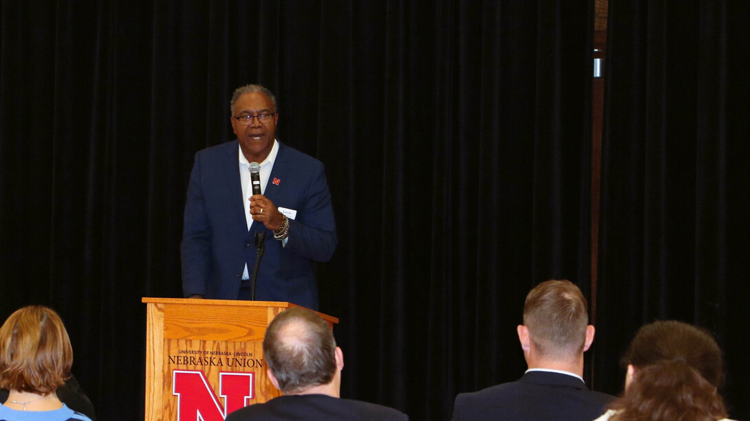 A Black man in a blue suit and tie speaks at a podium in front of an audience.