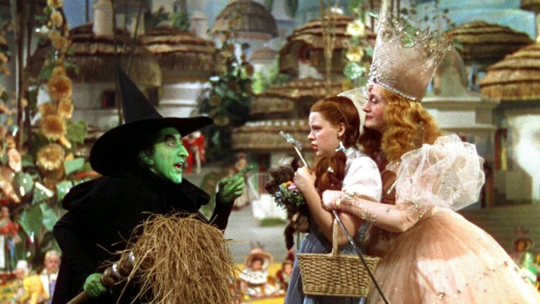 Dorothy, a white woman with long red hair, cowers in the arms of Glenda the Good Witch, a white woman in a pink dress and large tiara, in front of the Wicked Witch of the West, a green-skinned woman in a black jacket and pointy hat.