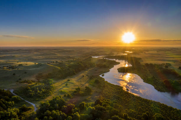 Green flat land with a river and shrubbery and trees and a sunset in the background