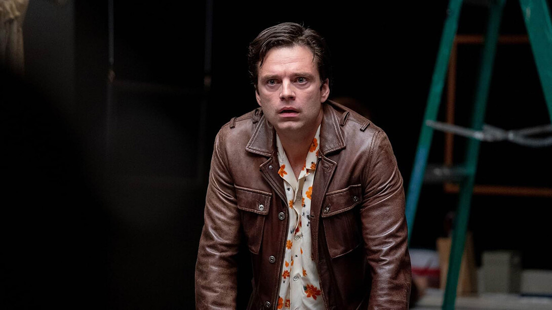 Sebastian Stan, white, with short brown hair, stands in a floral shirt and brown leather jacket.