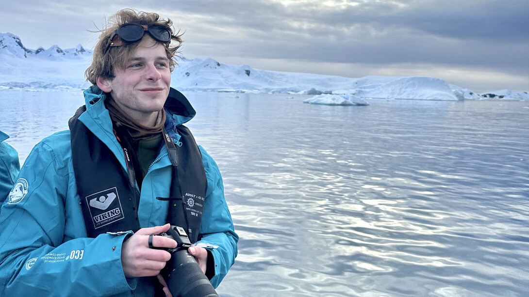 Luke McDermott, a white man with brown hair, stands before a large arctic ocean and icebergs in a blue jacket, holding a camera.
