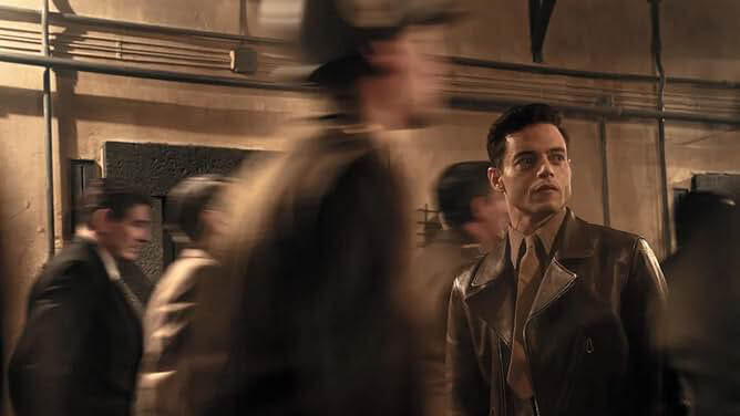 A white man in a brown leather trench coat and a suit and tie, with short brown hair, is surrounded by figures blurring past the camera.