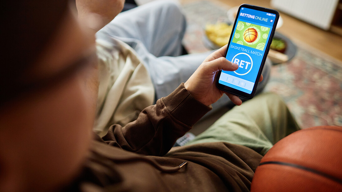 A young man holds a basketball as they sit on a couch while making a bet on a game via a cellphone.