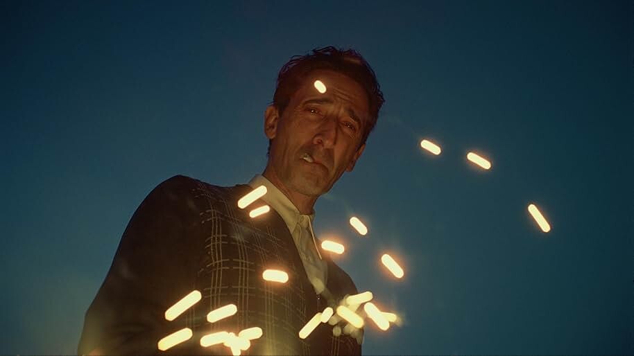 Adrien Brody starts as László Toth in "The Brutalist," standing, looking over the camera in a suit with a blue sky above him, smoking a cigarette.