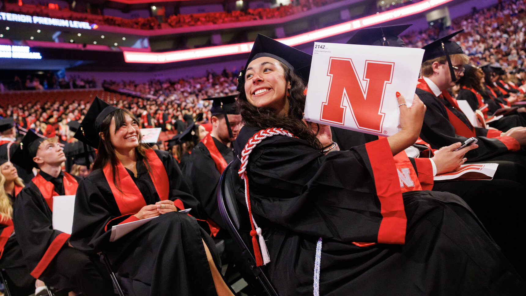 A female graduate, seated among other graduates on the floor of Pinnacle Bank Arena, smiles as she holds up her newly earned degree for family in the crowd to see.