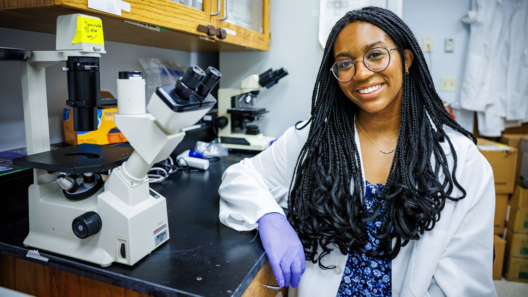 Laila Doyle, a senior nutritional science and dietetics major at the University of Nebraska–Lincoln, poses next to two microscopes in a lab. She is wearing a white labcoat over a blue floral dress, with purple protective gloves and glasses.